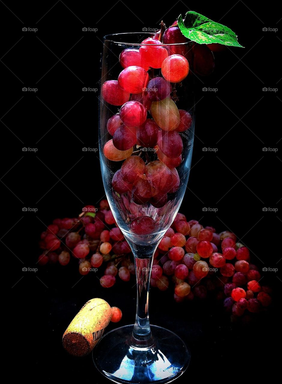 Fruit. In the glass is a bunch of pink grapes with a green leaf. Next to the glass is a bunch of pink grapes and a wooden cork from a bottle of wine. Black background