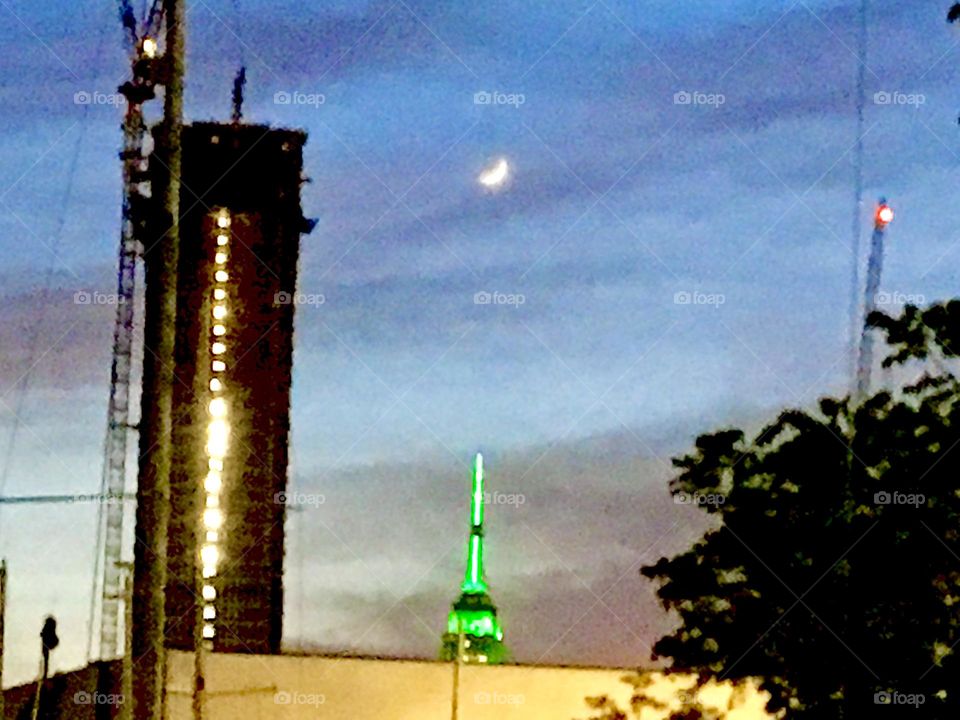 A twilight time view of the illuminated Empire State Building rooftop, a waxing moon in the sky and the dark silhouette of a high rise with lit up windows that reflect the last rays of the setting sun. 2021. Hypnotic Productions