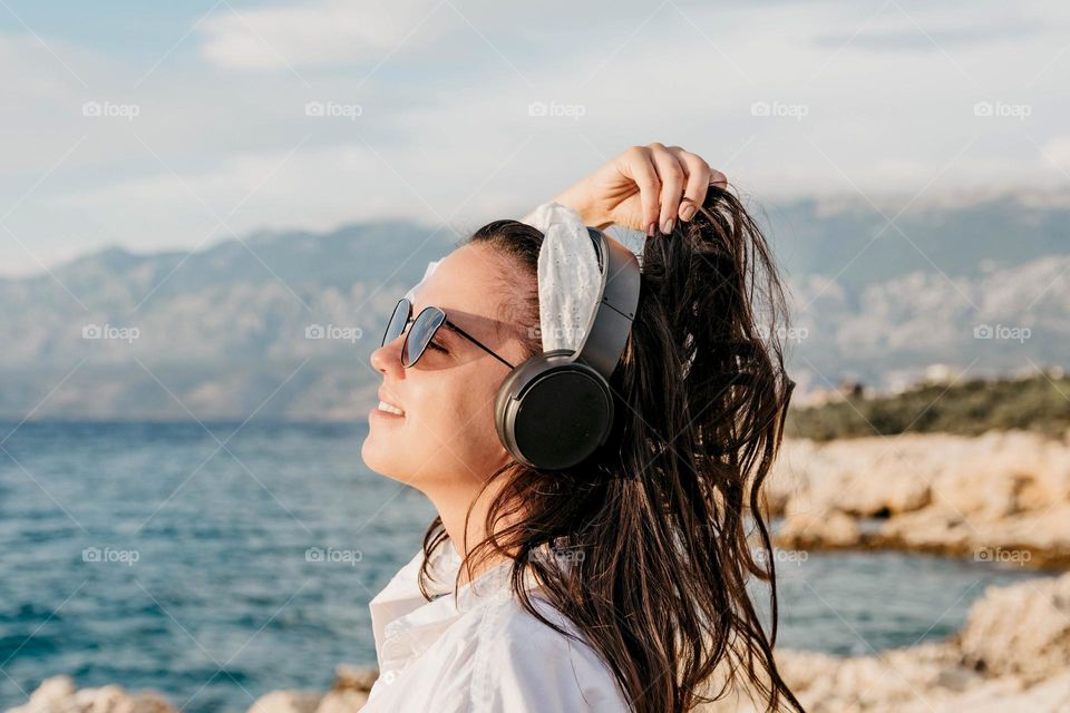 Beautiful girl with headphones on a beach. Happy woman weaing sunglasses, holding hair, enjoying on coast by sear