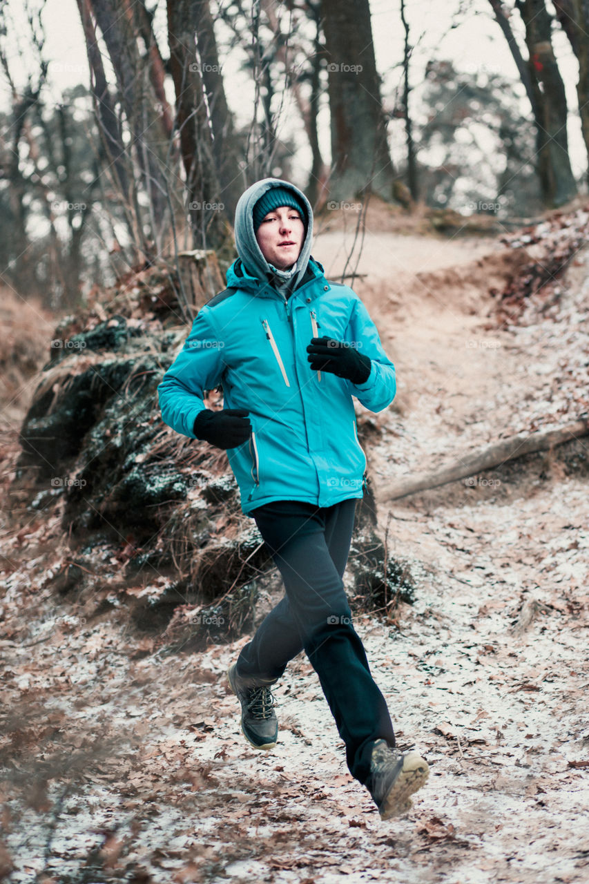 Young man running outdoors during workout in a forest among leafless trees on cold freeze winter day. Boy is wearing sport clothes