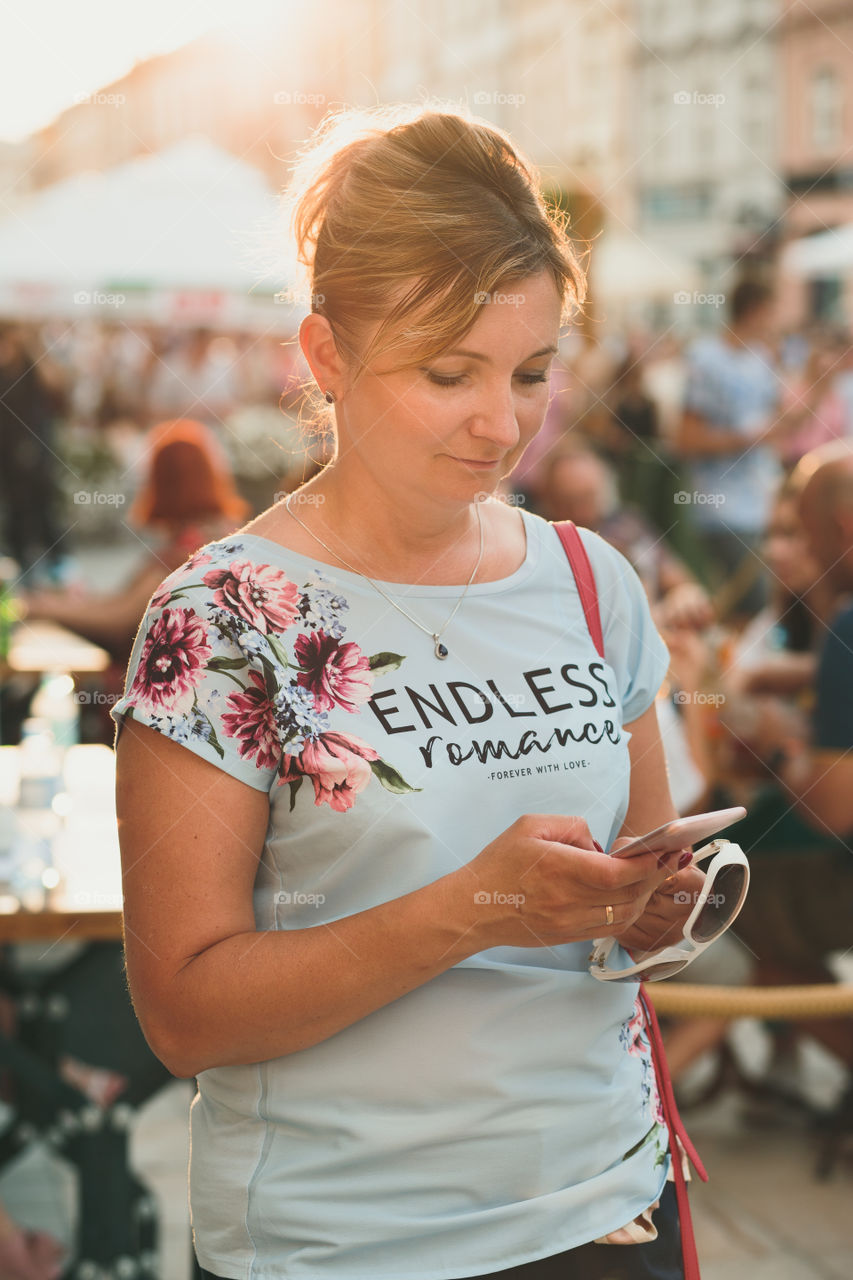 Smiling woman using mobile phone standing in the crowded center of town, backlighted by sun, wearing summer clothes