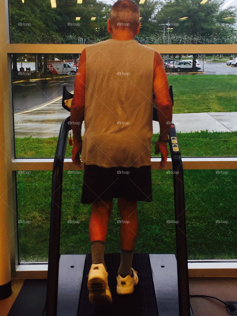 An elderly man at the gym walking on a treadmill on a rainy day. View of the outdoors and rain on the Windows 