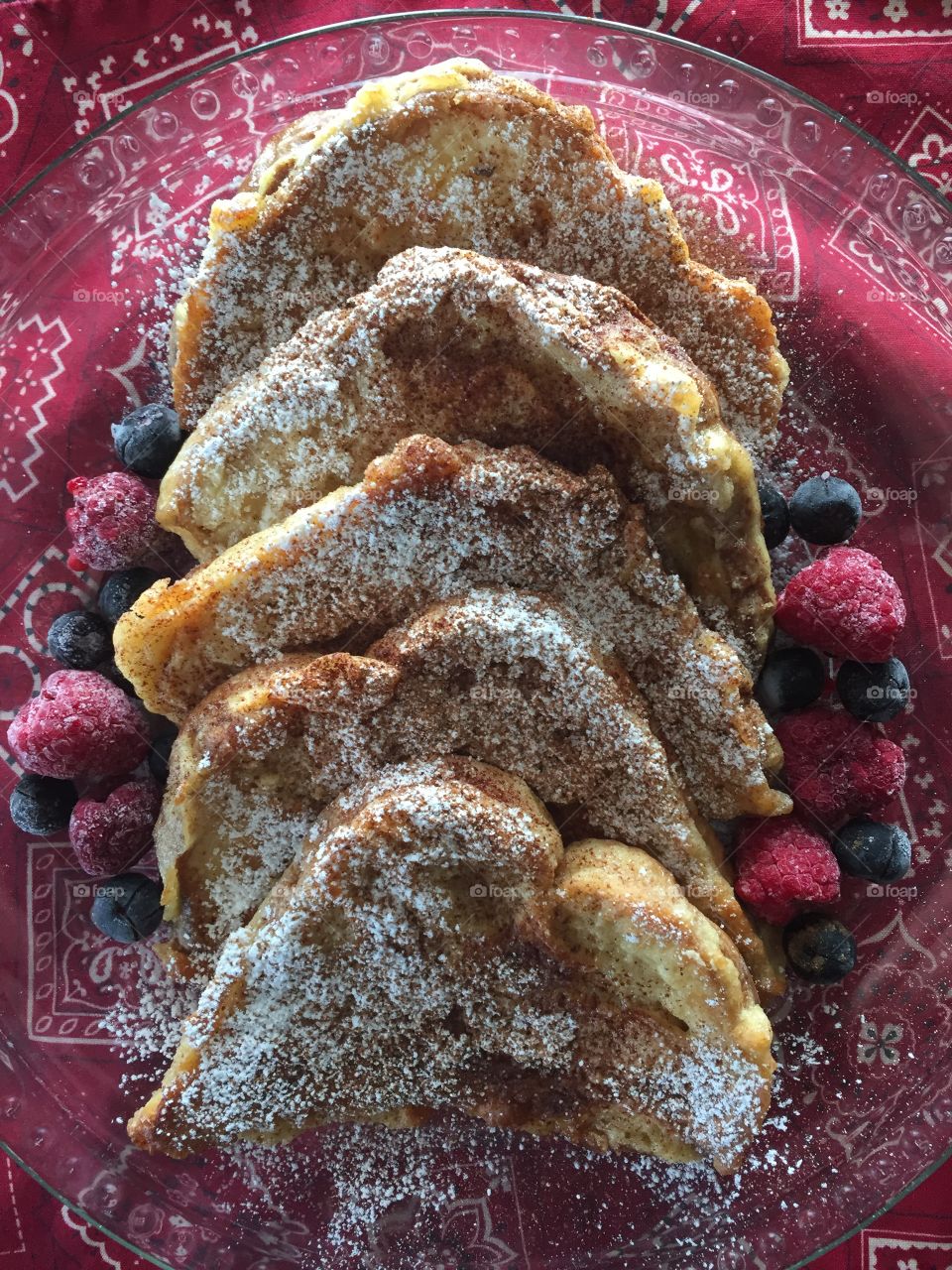 French toast on a clear glass plate, sprinkled with cinnamon and sugar, served with blueberries raspberries on a red handkerchief-patterned placemat