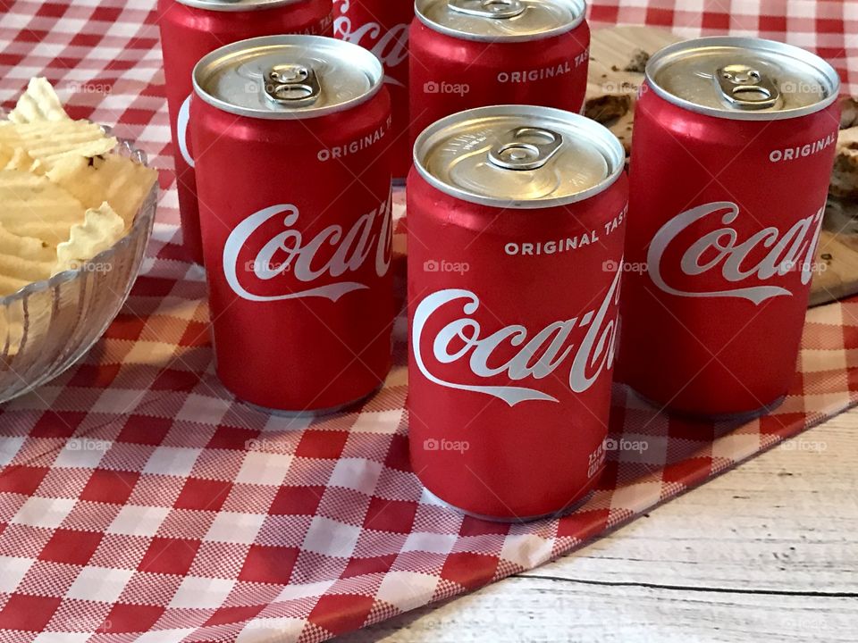 Cans of Coca-Cola on a red and white checkered background with barbecue food and potato chips 
