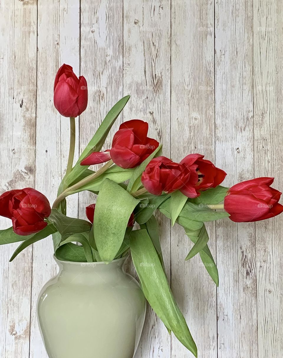 A bunch of red tulips in a green vase, against a pale wooden background 