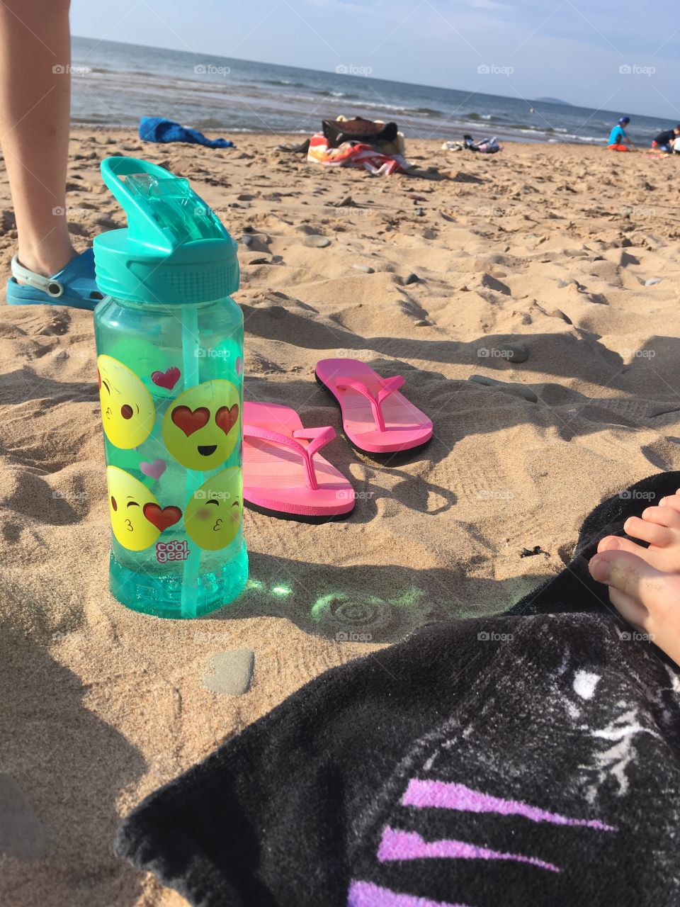 Beach day! Teal emoji water bottle, pink flip flops on sand. Bare feet on black towel. 