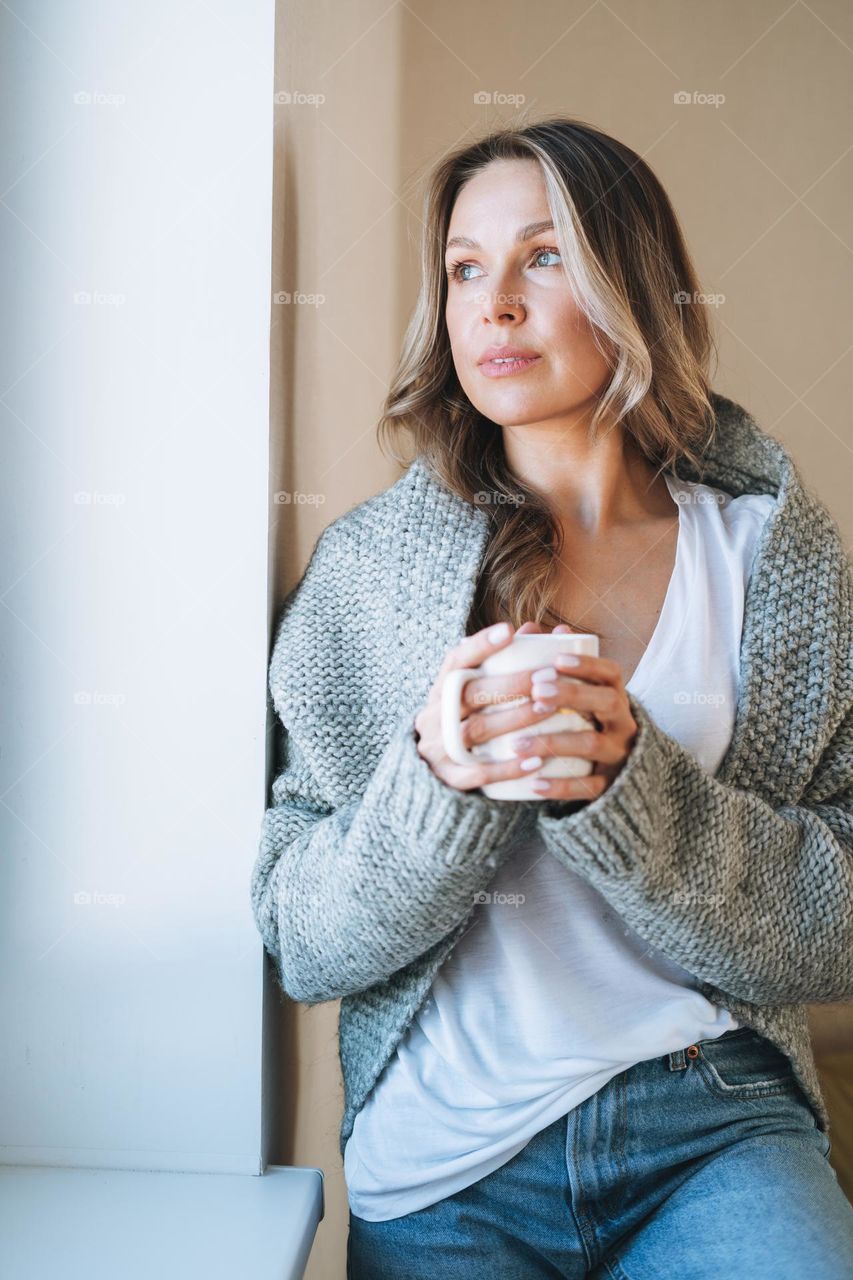 Young beautiful woman forty year with blonde long curly hair in cozy knitted grey sweater with cup of tea in hands in bright interior at home