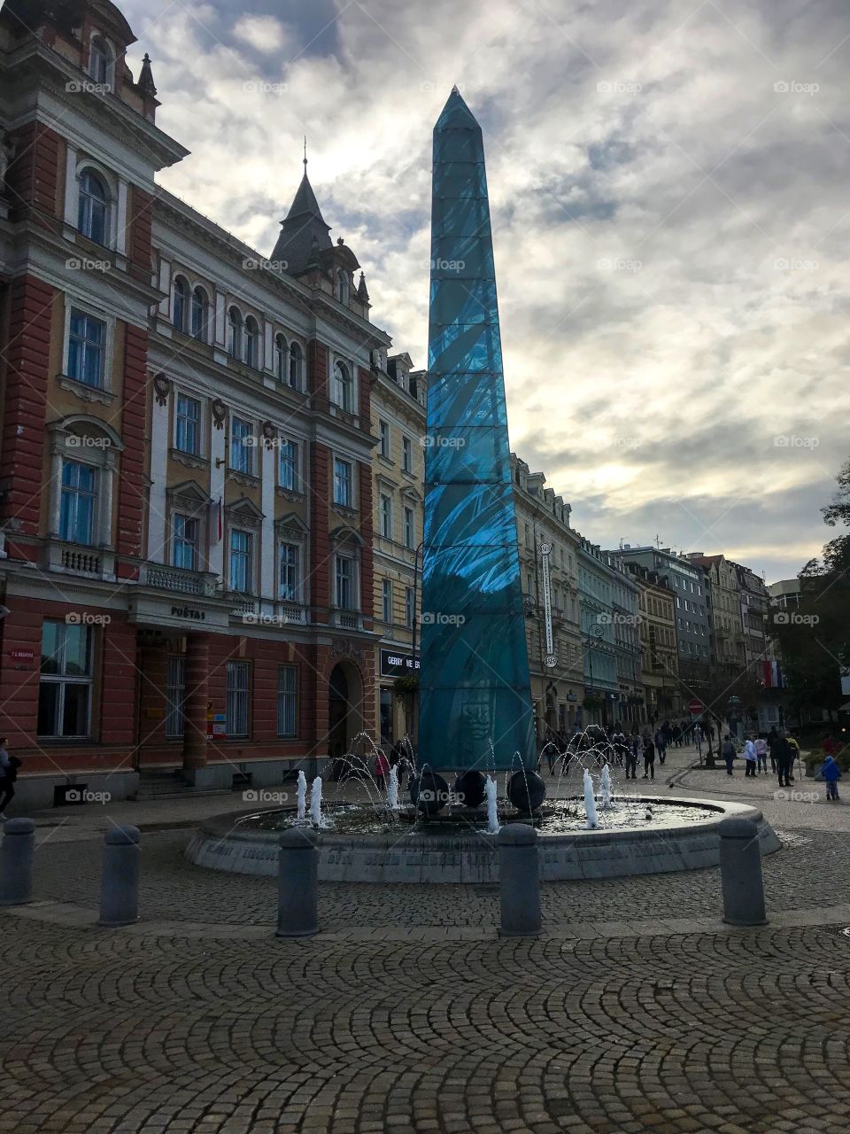  A pillar in the middle of the fountain , a series of buildings of different colors .There are many people walking in the distance, the sky was cloudy, with the golden glow 