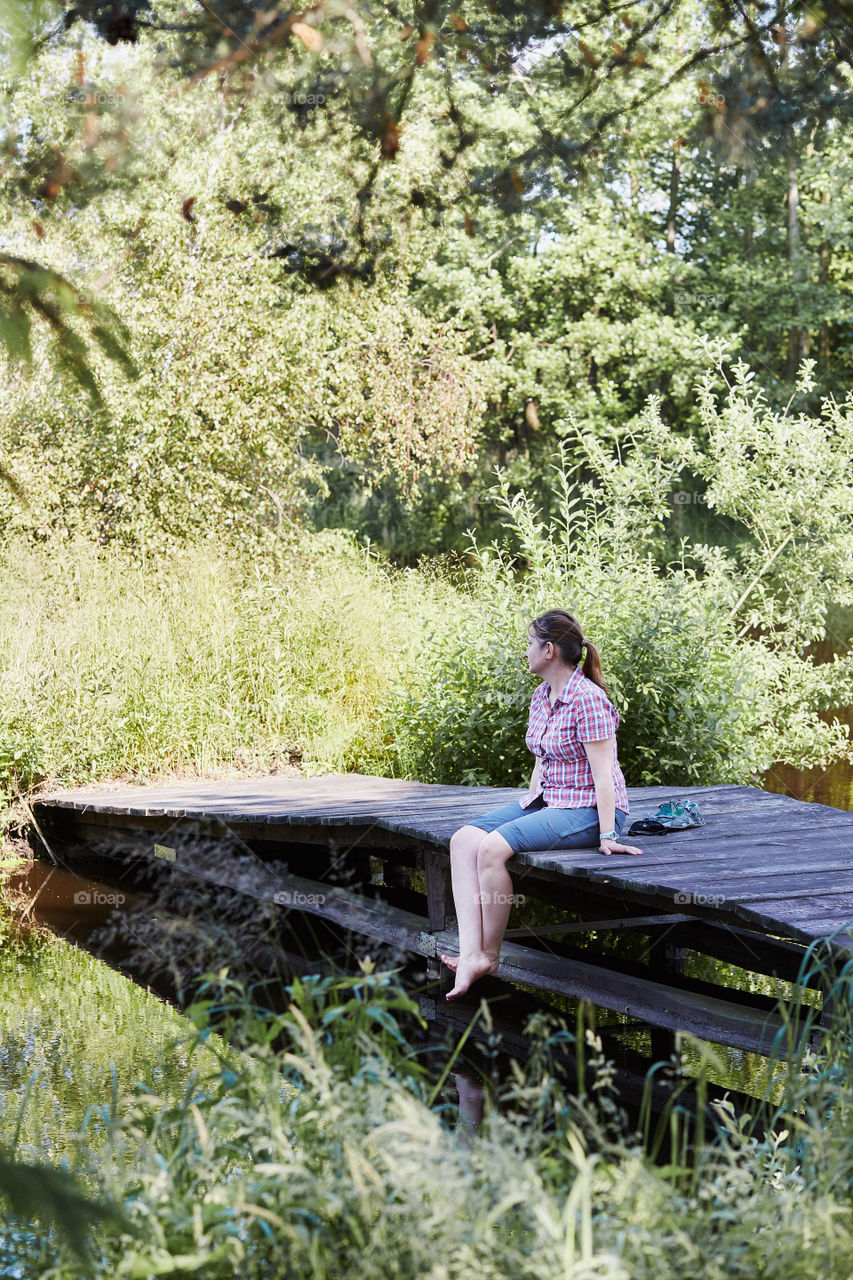 Woman sitting on a bridge over a lake, among the trees, close to nature, during summer vacations. Candid people, real moments, authentic situations