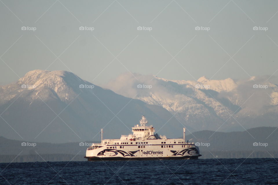 Shot of a BC ferry crossing the strait lit with the rare winter sunlight. The sea is indigo blue, the hills behind dark green, the mountains capped with fresh snow and small wisps of powdery clouds kiss the mountain peaks.
