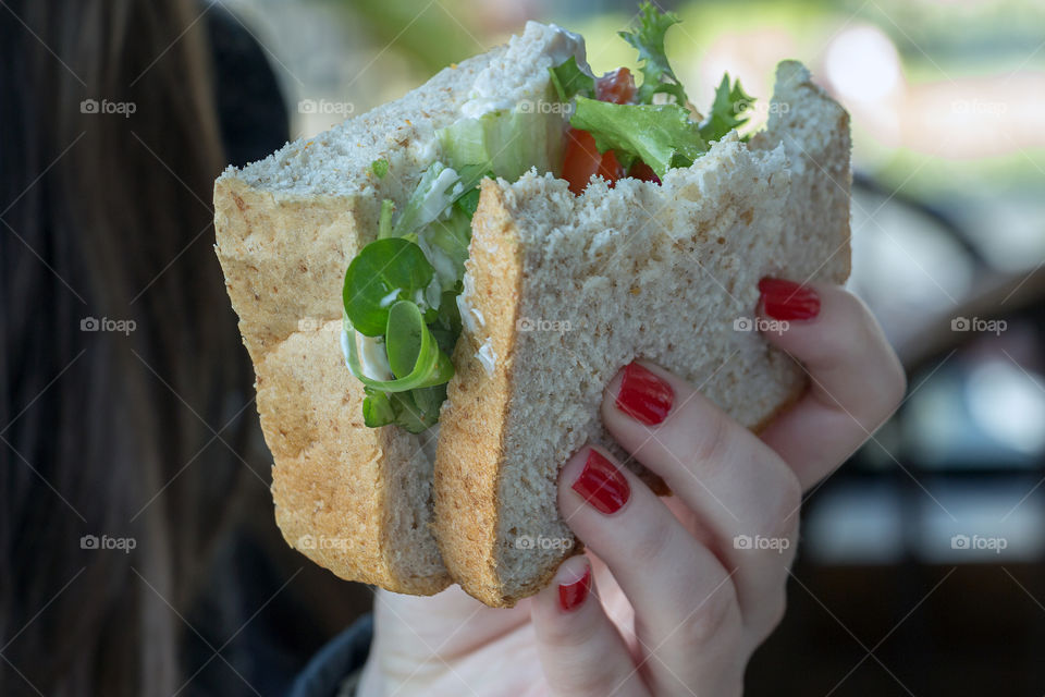 A girl’s hand holding a sandwich with vegetables, sliced cheese and meat between sliced of bread