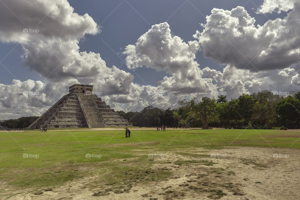 Panorama with the Pyramid of the archaeological complex of Chichen Itza in Mexico surrounded by natural vegetation under a sky with sheepish clouds and some tourists who admire it ecstatic.