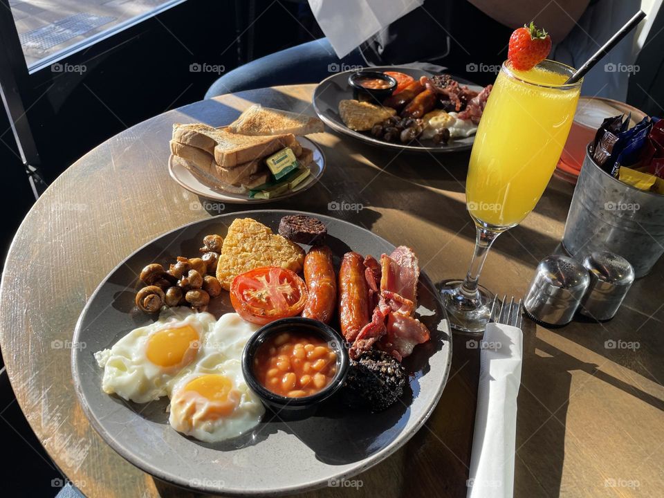 The table is set with a hearty Irish breakfast. Next to the plate sits a glass of sparkling mimosa, its bright orange juice and champagne bubbles offering a refreshing contrast to the rich and savory meal.