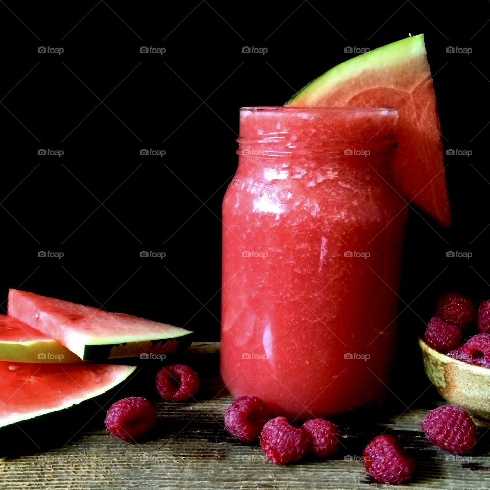 Watermelon smoothie in glass with watermelon wedges and fresh raspberries on rustic wood table.