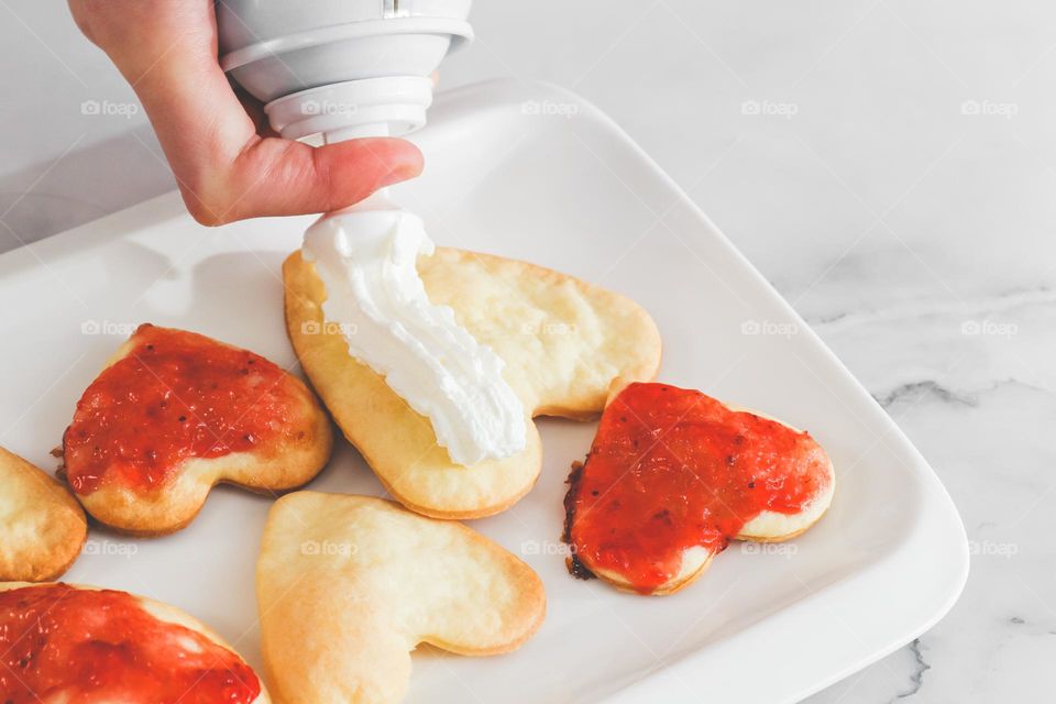 The hand of a caucasian girl adrostok squeezes whipped cream from a balloon onto homemade cookies with jam in the shape of a heart in a boyud, standing at a moamer table, side view close-up.