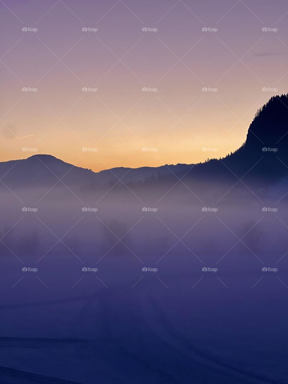 A dreamy winter sunset where golden light melts into misty purple mountains. The soft fog cloaks the snowy landscape in mystery, while the last light of day paints the sky in shades of serenity. A peaceful moment captured in nature’s quiet magic.