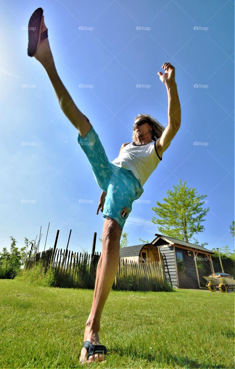 dynamic portrait from a man wearing sunglassed kicking his leg high up in the air on a warm summers day with blue sky wide angle frog perspective picture