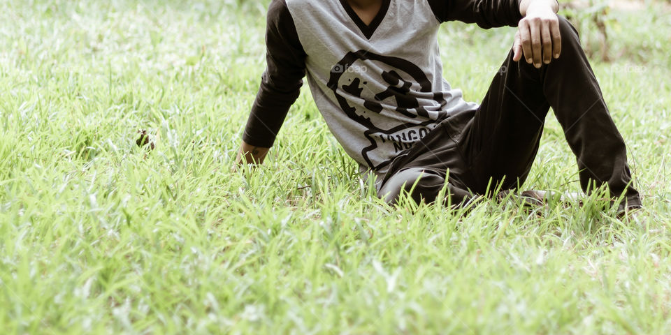 Close up Cropped image of young man in casual clothing sitting over green grass in the park at morning time of day. Front view. Concept of spending leisure time in open air surrounded by nature. The human body surrounded by beauty in nature concept.