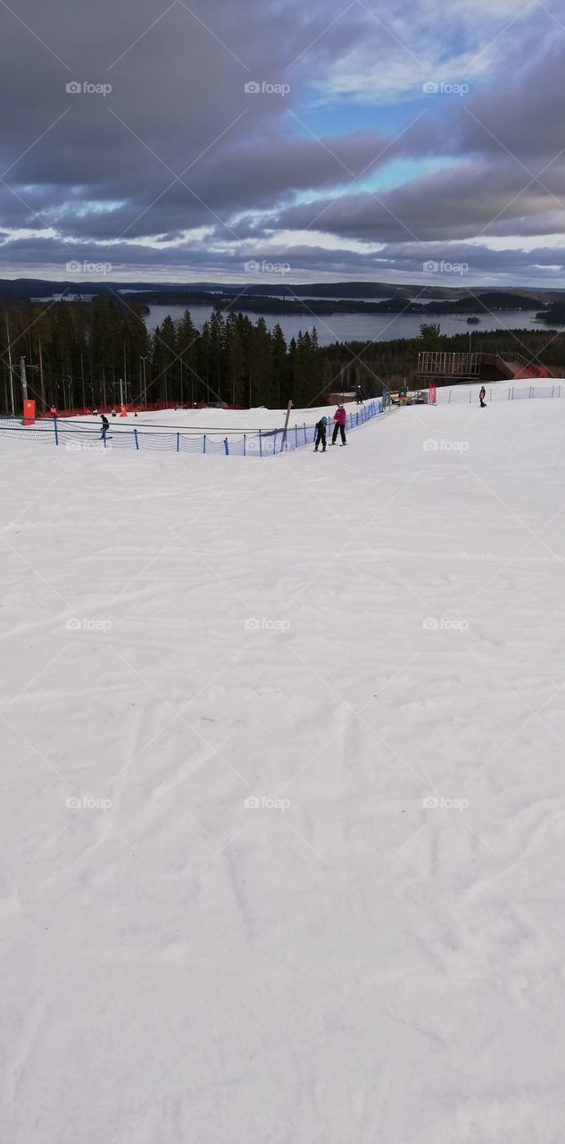 People on the ski slope skiing down and lifting up on the bright snow. In the background a lake, an island and forests.