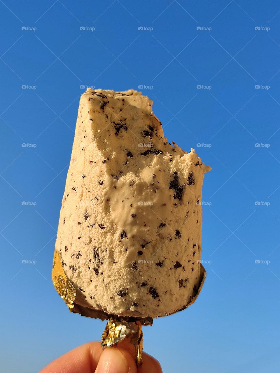 Sunny day.  Against the background of the blue sky, a woman's hand holds an ice-cream with chocolate chips.  A piece was bitten off from the ice cream.  Temptation and joy