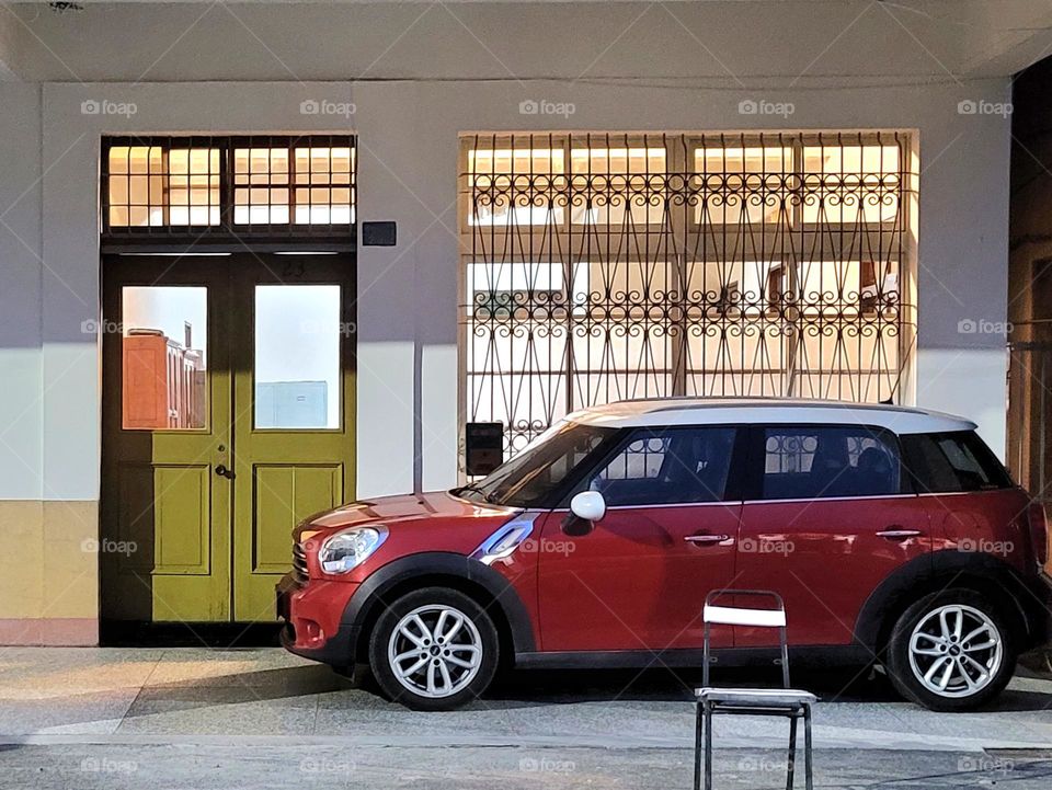 A car with a white roof and a red body is parked in front of a building with iron windows showing yellow light