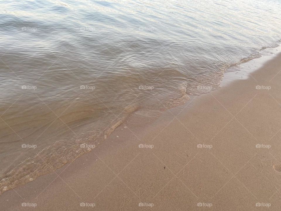 This image captures a close-up view of gentle waves lapping against a sandy beach. The water appears calm, and the shoreline is smooth with small bubbles forming at the edge where the water meets the sand, a peaceful and serene beach scene.