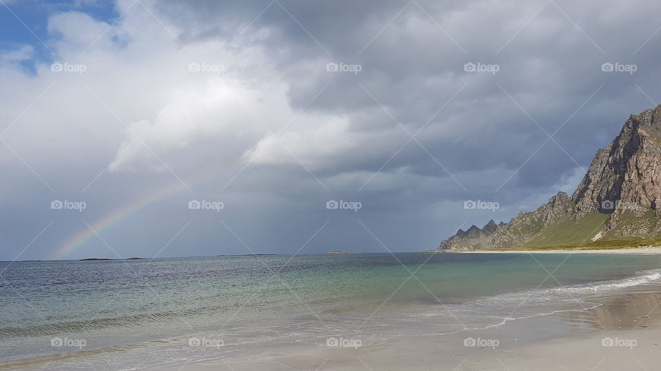 A beach near Bleik with a rainbow in background and cloudy sky, Arctic Norway.