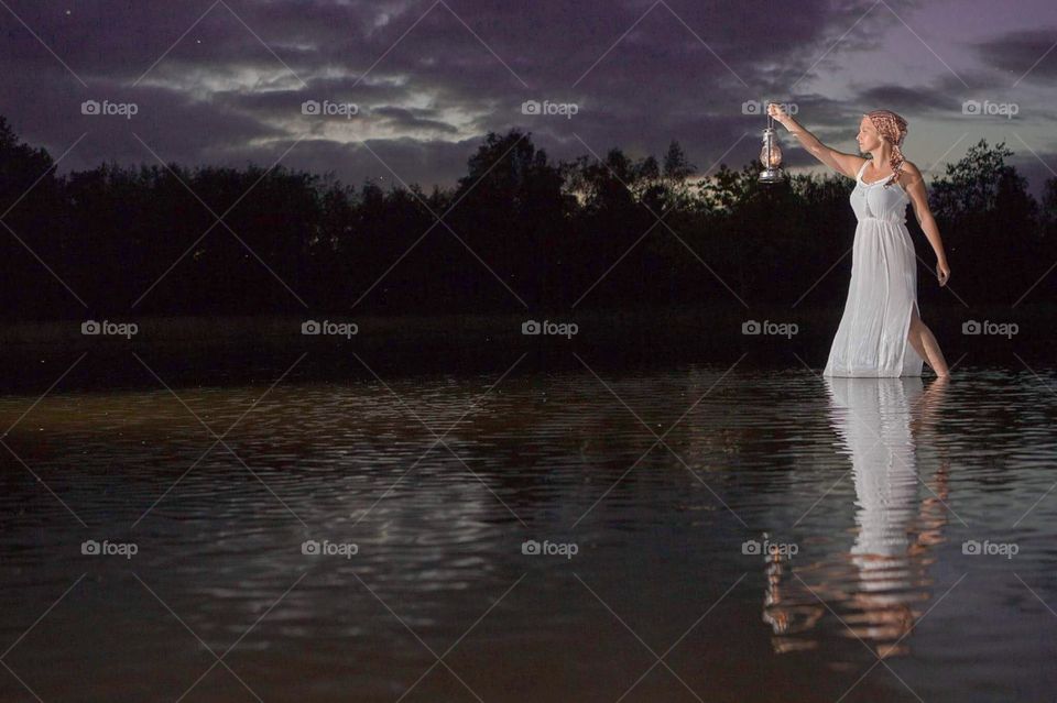 Beautiful young blonde woman standing in the water of a lake around Dusk, sunset, wearing a long white dress and holding a vintage lamp 