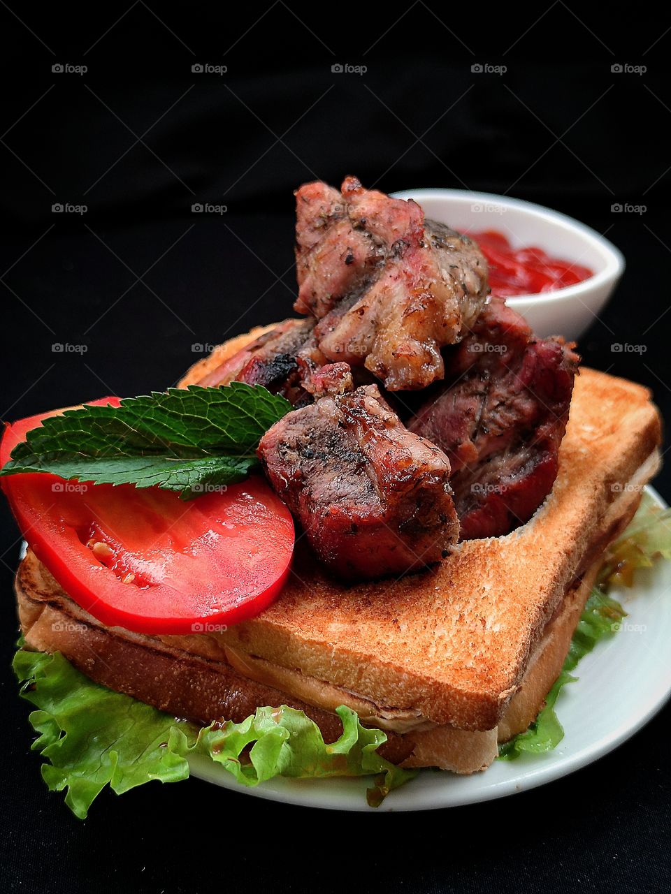 on a white plate: green lettuce leaves, croutons with pieces of fried meat, a piece of red tomato with a green mint leaf on it. In the background is a white gravy boat with red ketchup. Black background