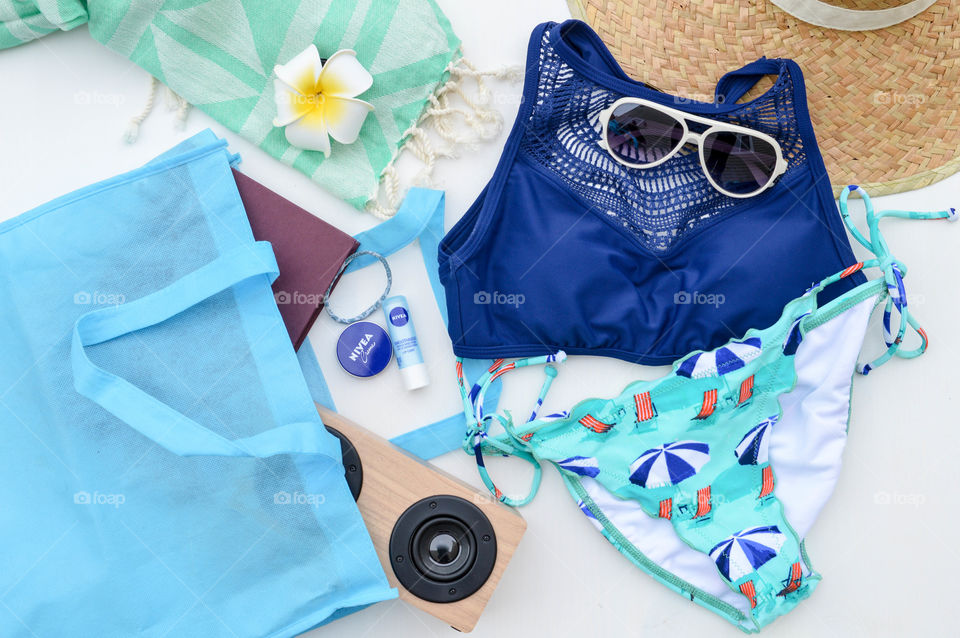 Contents of summer bag laid out on a white table consisting of a swimsuit, Nivea cream, Nivea lip care, a book, sunglasses,  hair tie, speakers and towel