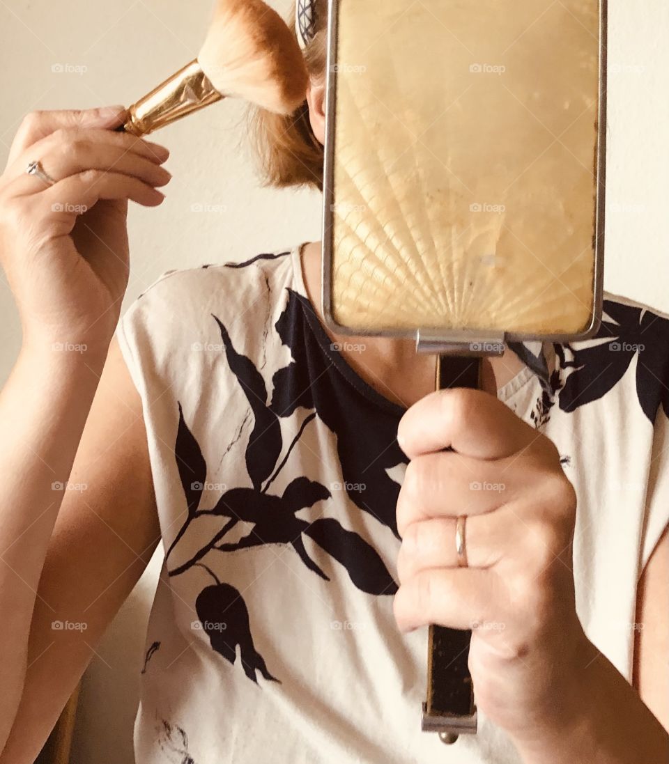 Woman behind art deco hand mirror held in one hand and a makeup brush in the right hand