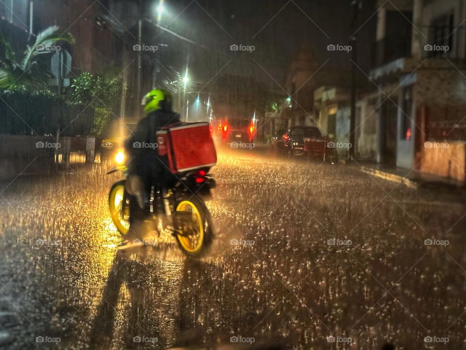 Man riding a food delivery motorcycle crossing a street on the rain at midnight, while a car illuminates him.