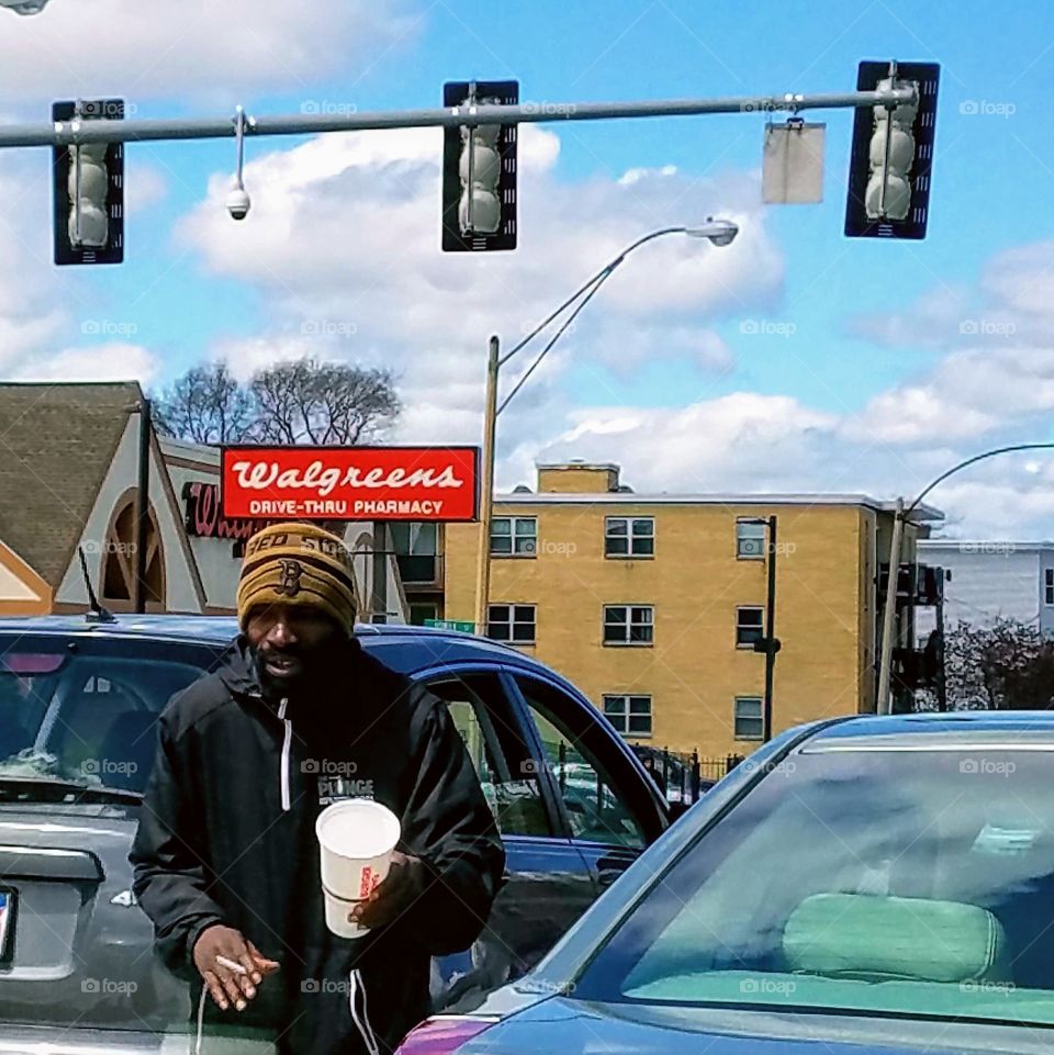Panhandling in City traffic, cars stopped at lights overhead. He's  carrying a cup for drivers to put money into. Knit hat on mans head, cold weather out.