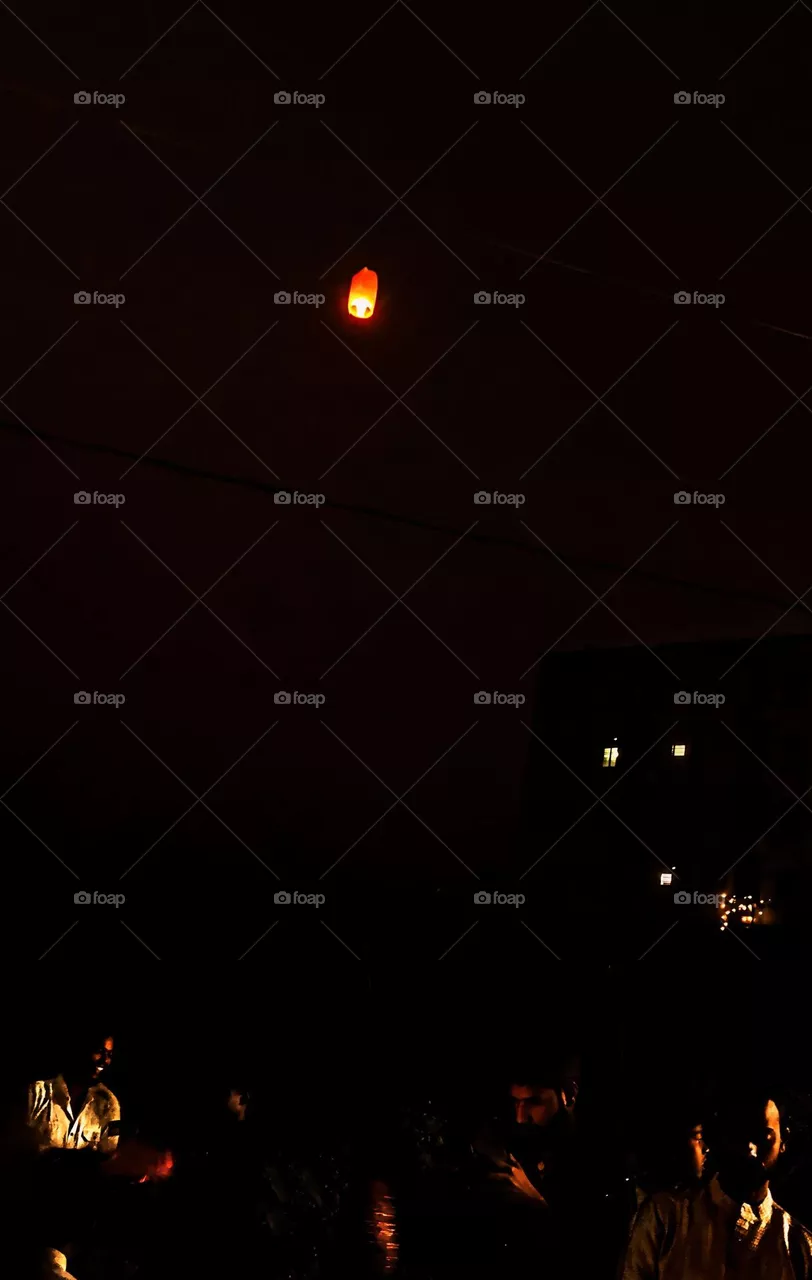 A glowing lantern ascends into a dark night sky, casting a warm light over silhouetted figures and buildings, capturing a serene moment of celebration and unity.