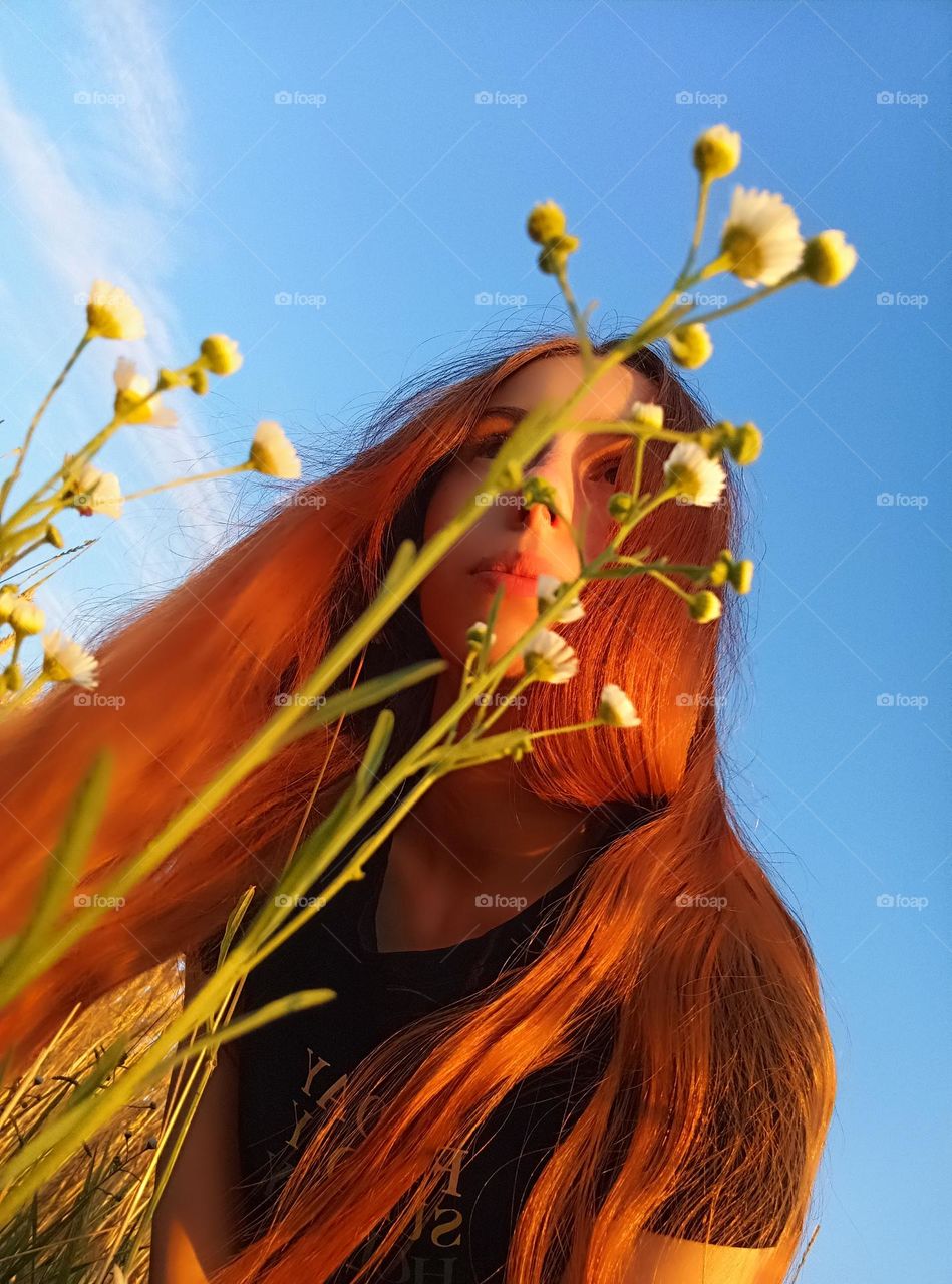 It is a photo of a young woman in the field. She has red (ginger) hair. She has brown eyes. Erigeron annuus ( the annual fleabane, daisy fleabane,  eastern daisy fleabane). Sky is blue.