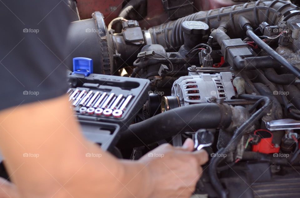 A man is working hard with car repairs to  his engine with a variety of tools as he attempts to install an alternator.