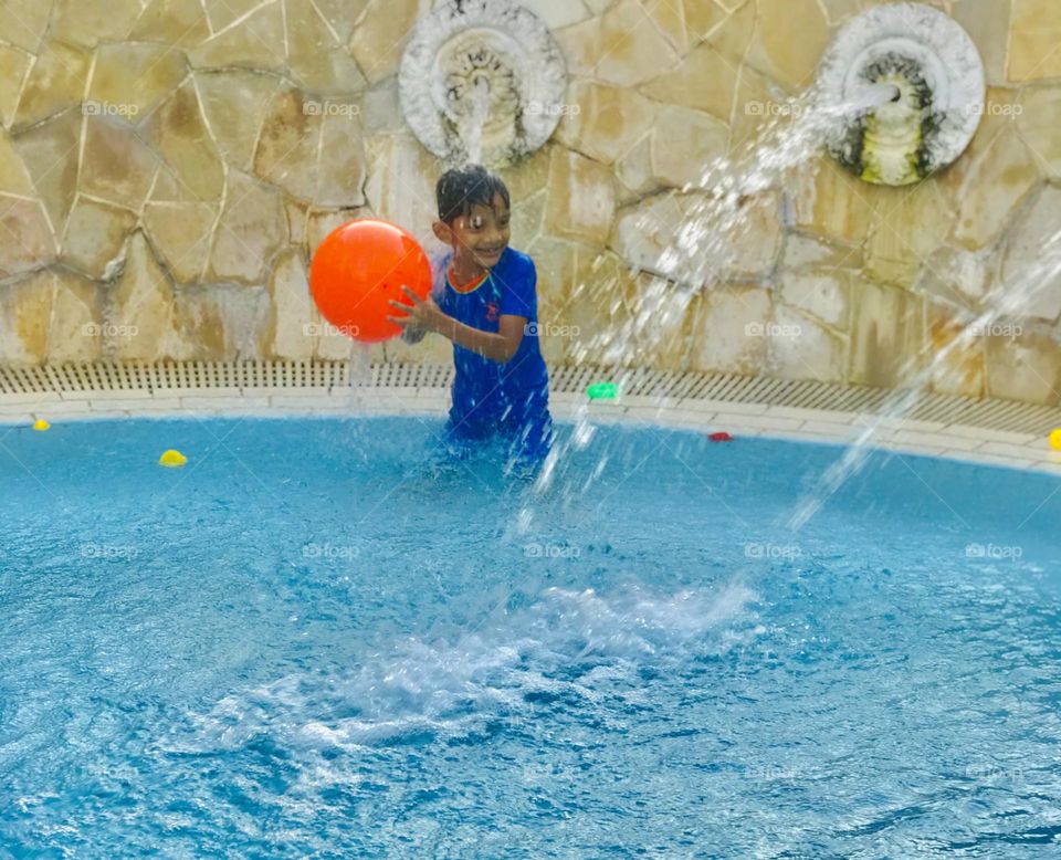 A boy enjoying a lot at swimming pool. He stands under waterfall and playing with orange boll and ducks and he has a great smile 😊