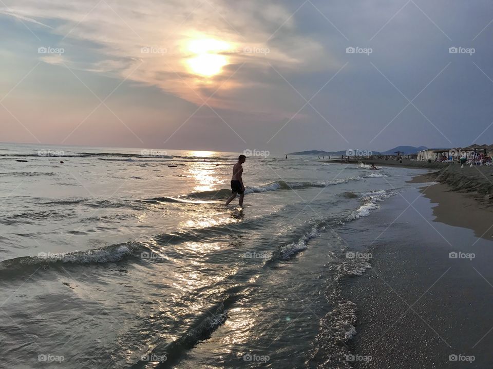 A man walking through shallow water in the sea, enjoying his time by the ocean, during the sunset.