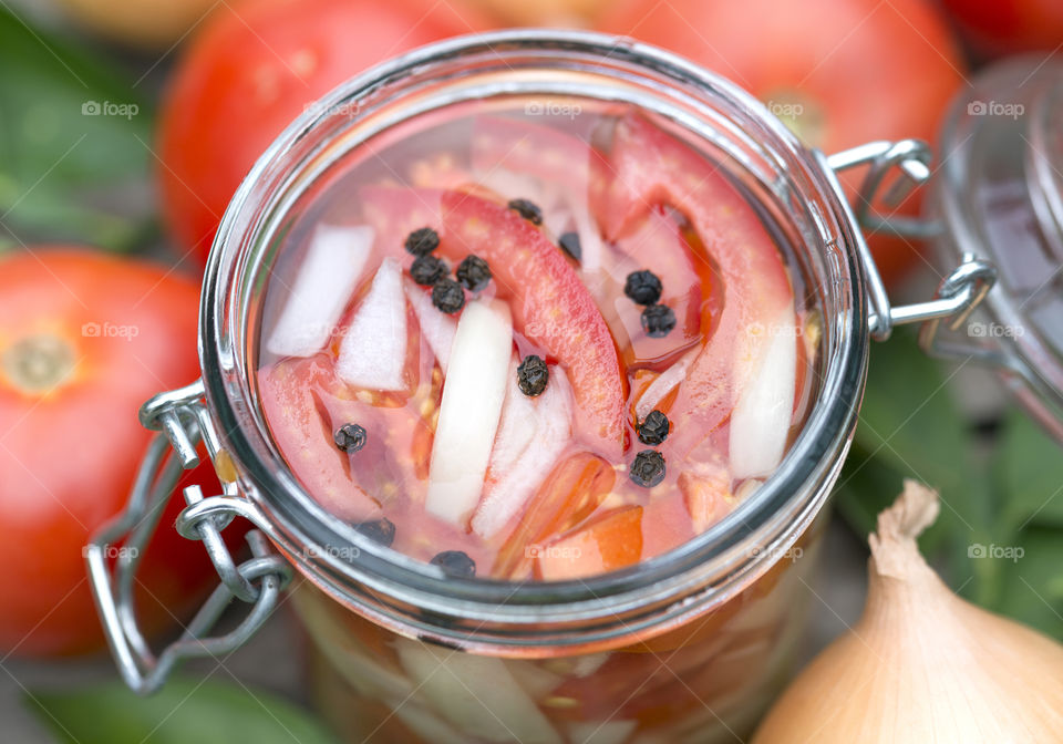 A closeup view of sliced red tomatoes, sliced onions and peppercorns in a glass jar.