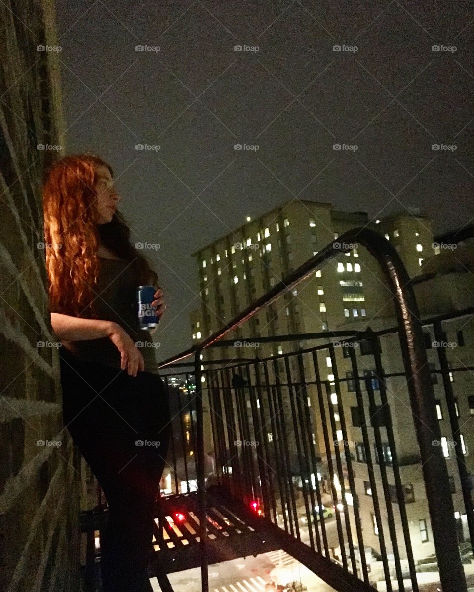 Red-headed girl holding a Bud Light leaning on a wall of aNew York City fire escape at night.