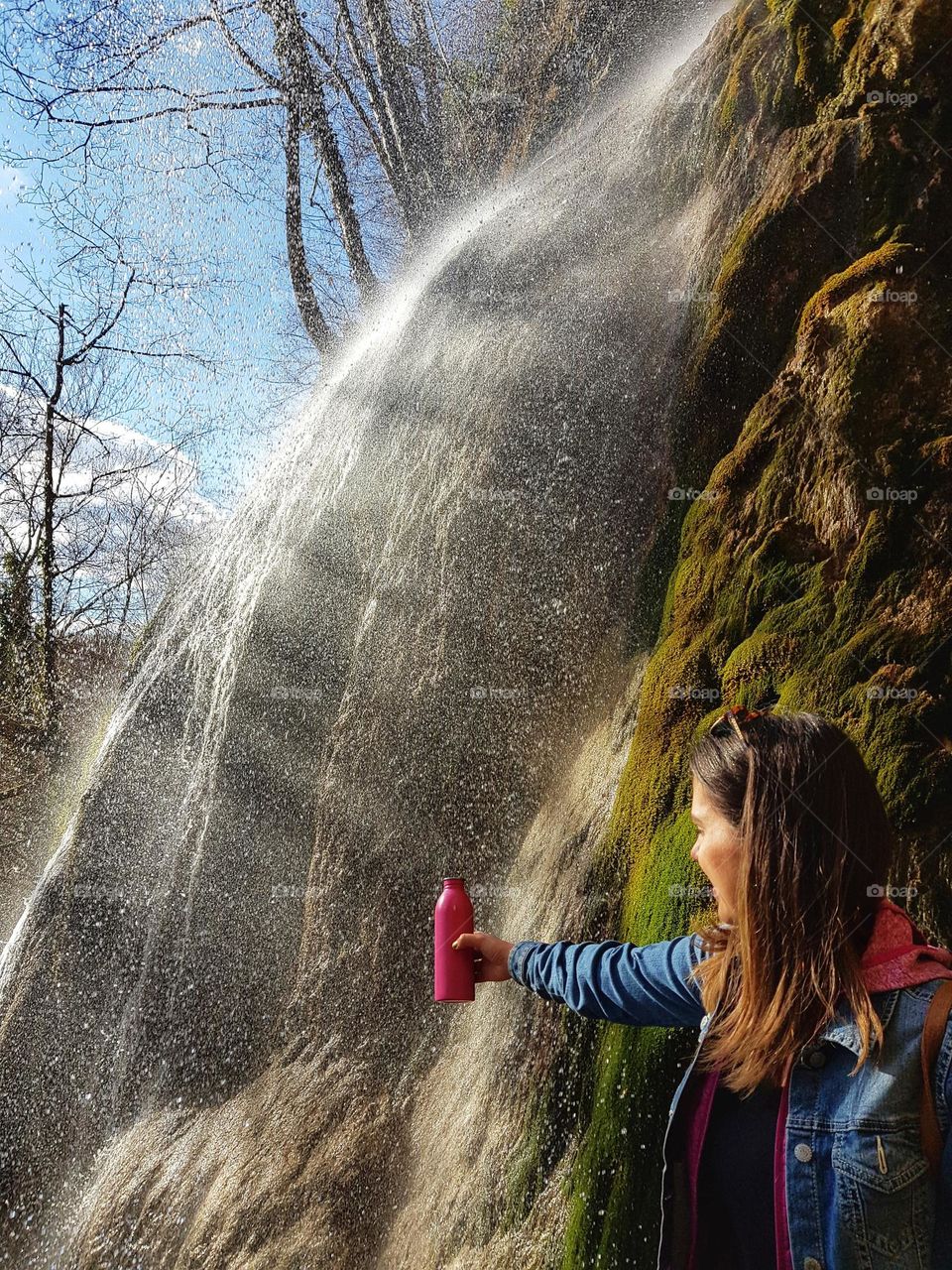 Happy young woman refilling water bottle at a waterfall on a sunny day in spring