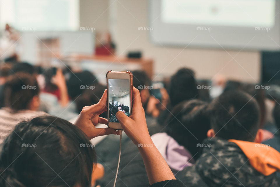 Rear view of girl taking picture of peaople using mobile phone. Visible smartphone screen