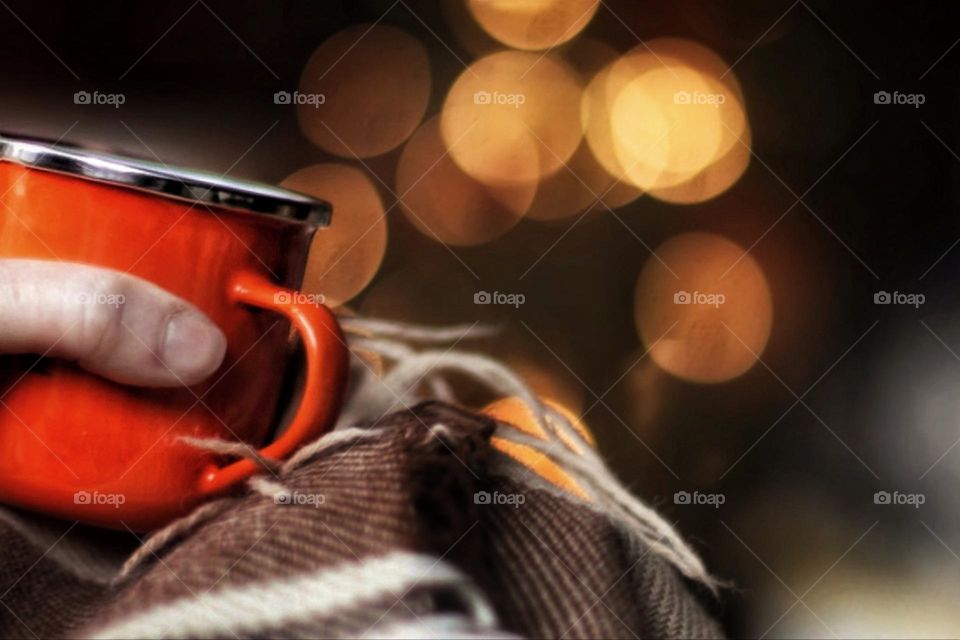 A hand holds a red cup on a brown woolen blanket against an orange illuminated background with bokeh effect 