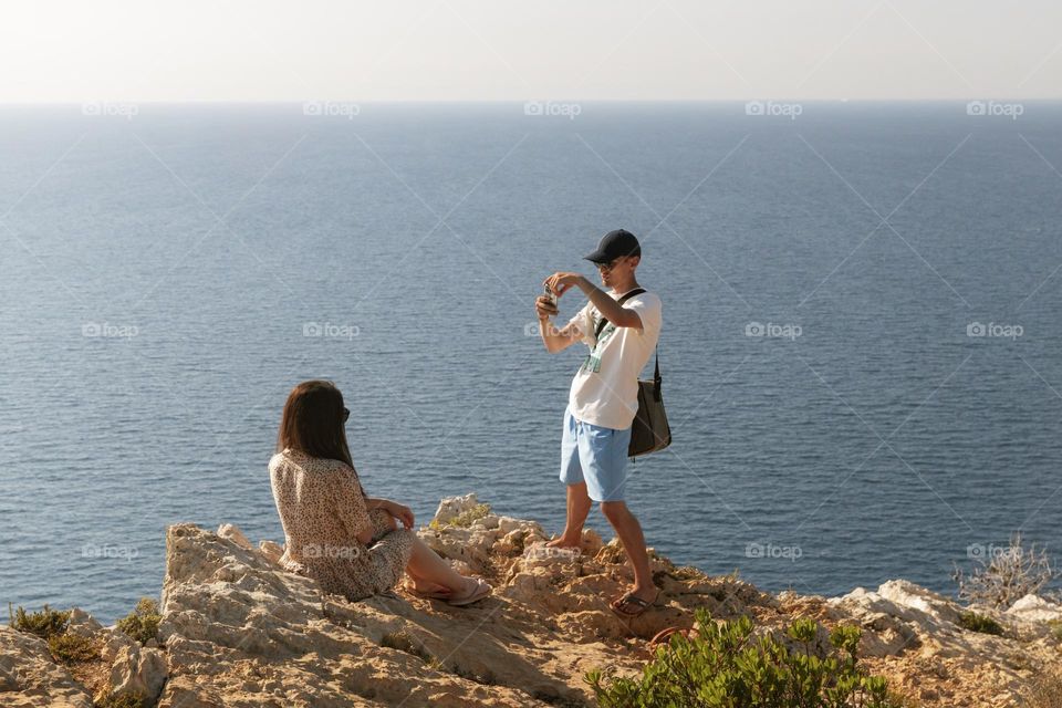 One young Caucasian guy takes a photo of his girlfriend sitting on a stone rock against the backdrop of the sea on a summer day on the island of Zakynthos in Greece, close-up side view.