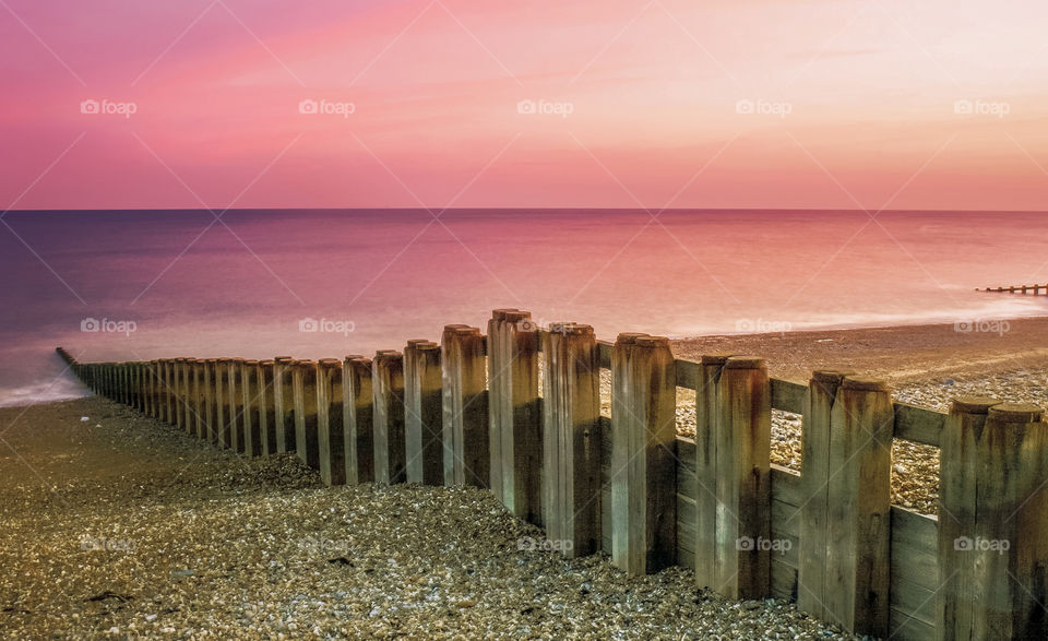 Dusky pink sky after sunset over the sea, in the foreground wooden beach groynes, cut across the pebbles and lead down to the shoreline 