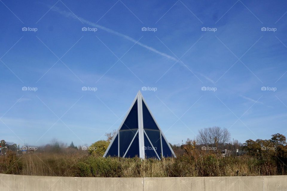 A triangular shaped building in the grass with a jet stream overhead.