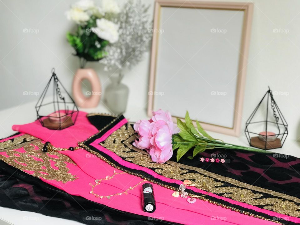 A pink colour saree and pink colour neck chain with pink beads and bracelet and hair pin along with flowers and candle everything pink in colour on white table with pink photo frame and flower vases .