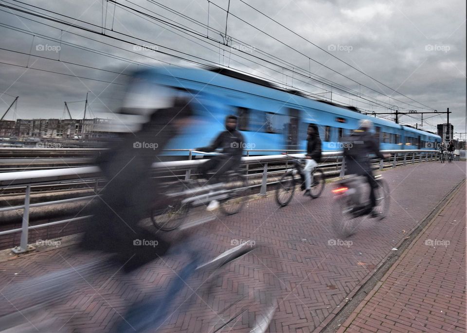Blue train passing by at full speed and cyclist cycling on the street in an urban environment with high voltage cables and clouds in the sky