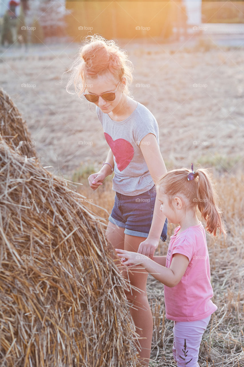 Sisters, teenage girl and her younger sister playing together with hay bale outdoors in the field in the countryside. Candid people, real moments, authentic situations