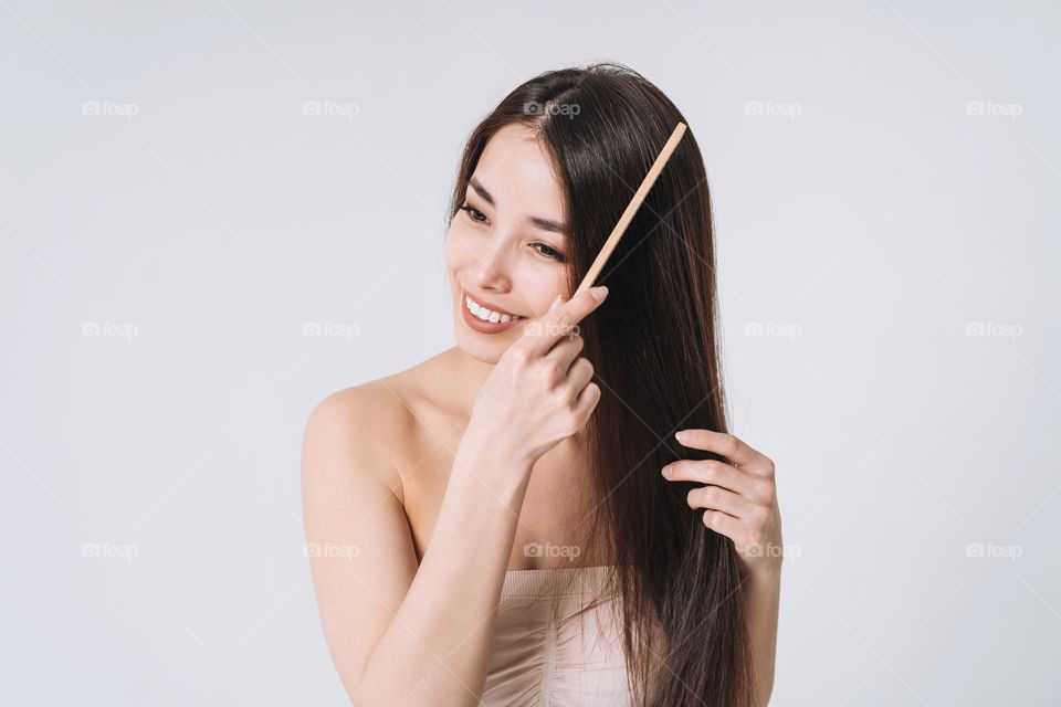 Beauty portrait of happy smiling asian woman with dark long hair combing wooden comb on white background isolated